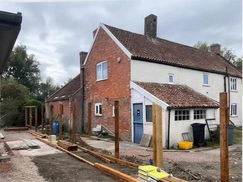 New fence posts set along a gravel drive beside a brick and render cottage, timber stacked ready