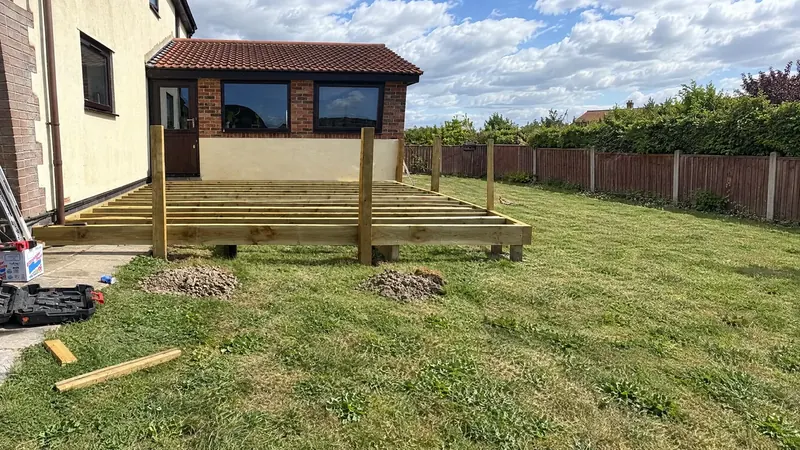 A raised timber deck frame on a garden lawn beside a house, posts and joists in place before the boards go down