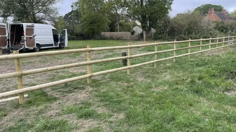 A long post and rail fence running across a field, trees beyond